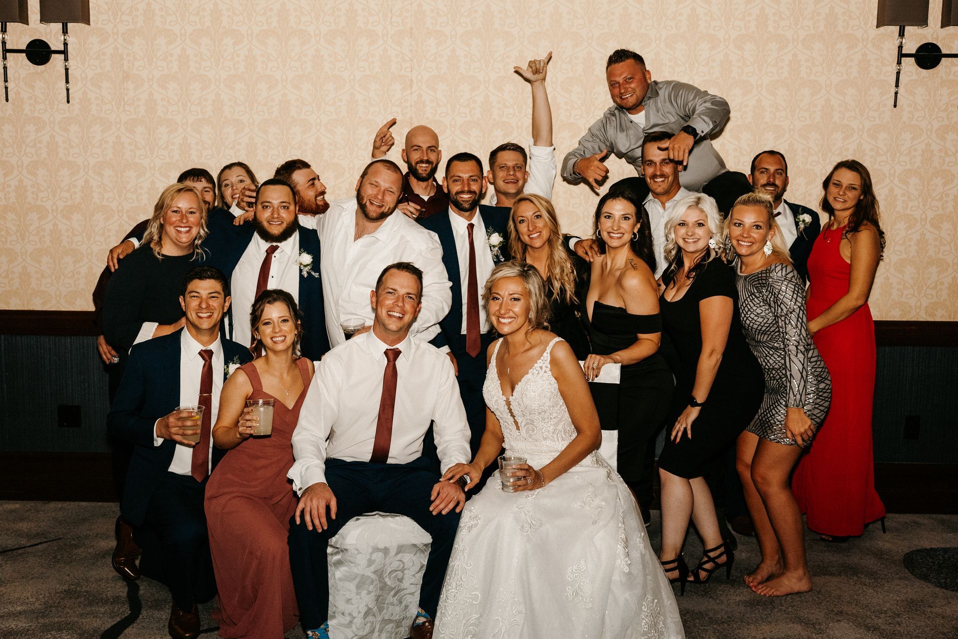 Wedding group photo with bride and groom, others smiling, some raising hands. Neutral setting, warm tones.