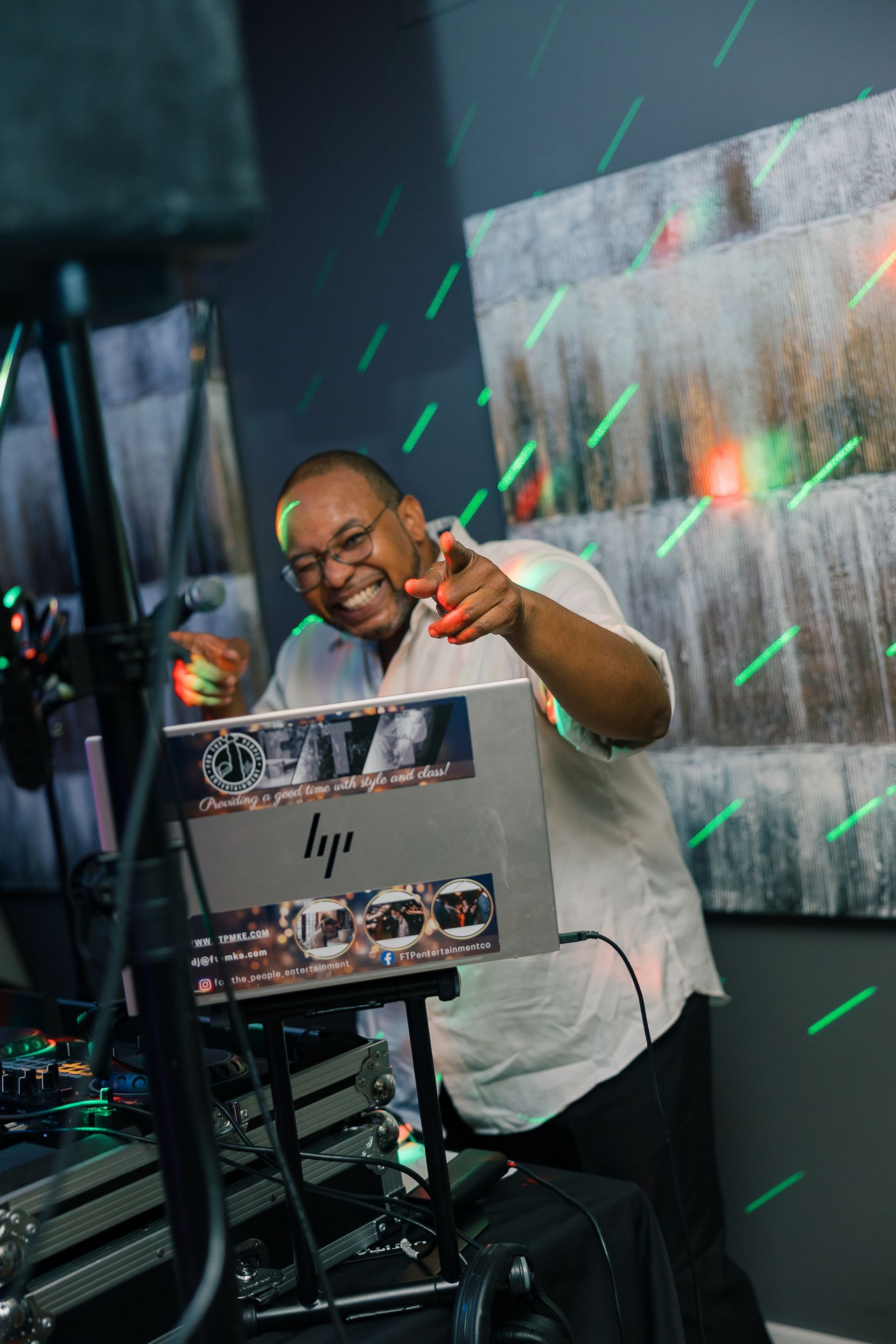 Man at DJ setup, pointing and smiling. Green and red party lights, gray wall in background.