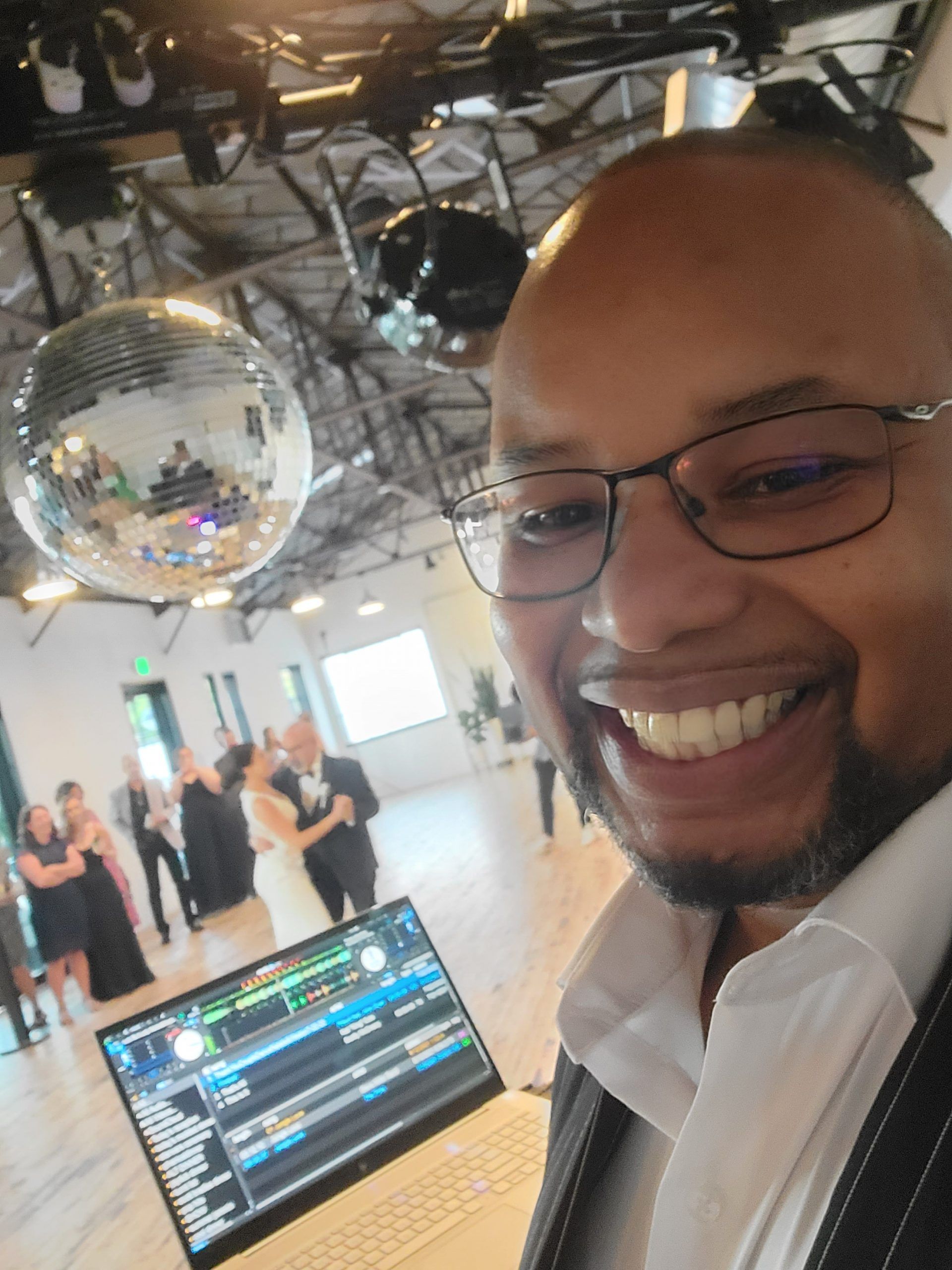 DJ smiling, holding laptop, disco ball in the background. Event setting, people dancing.