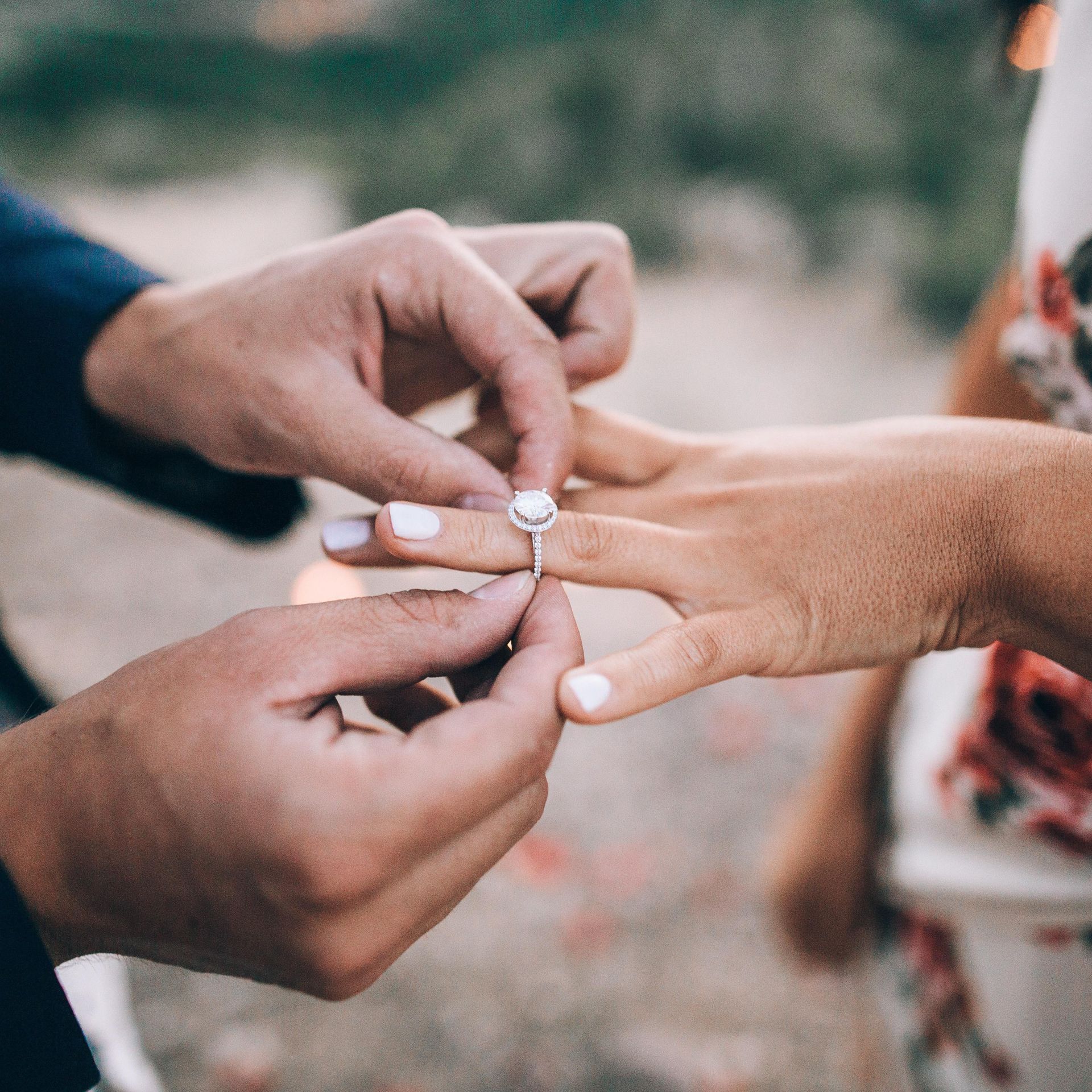 Man putting a ring on a woman's finger, outdoors. Focus on hands and ring; both have white nails.