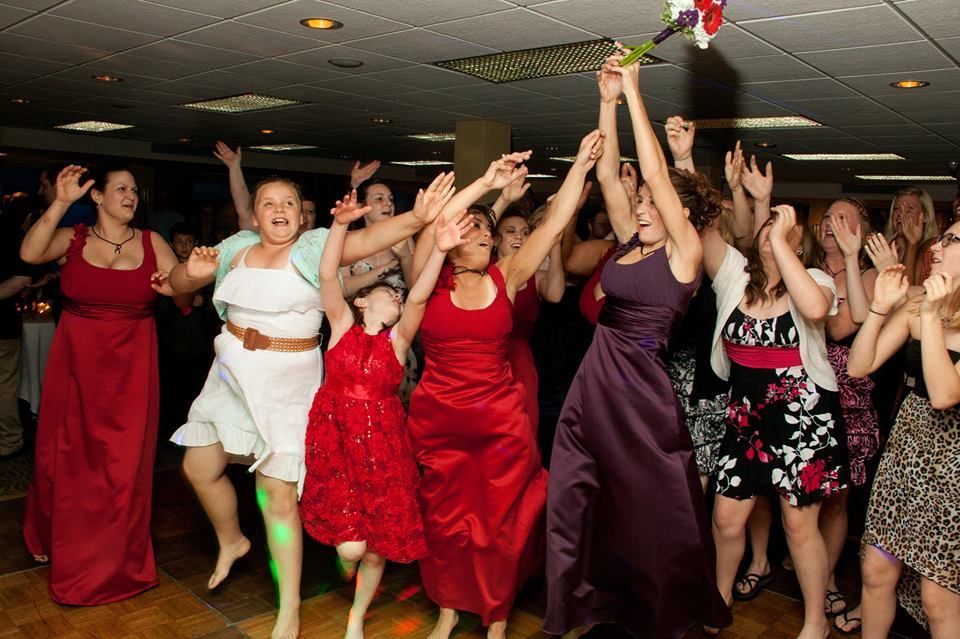 Women at a wedding reception reaching for a bouquet. They wear dresses in red, white, purple, and floral prints.