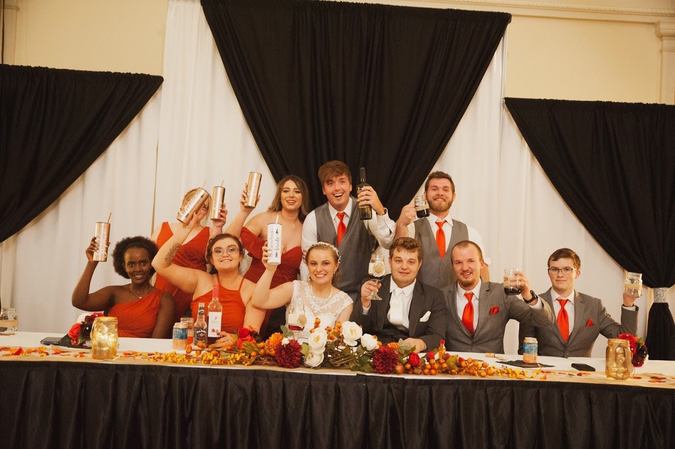 Wedding party cheers around a decorated table; red and white colors dominate.