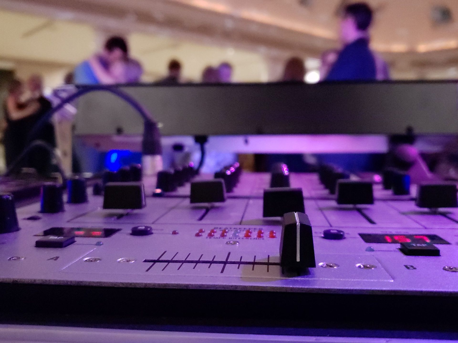 DJ mixer with knobs and faders, people dancing in background, purple and blue lighting.