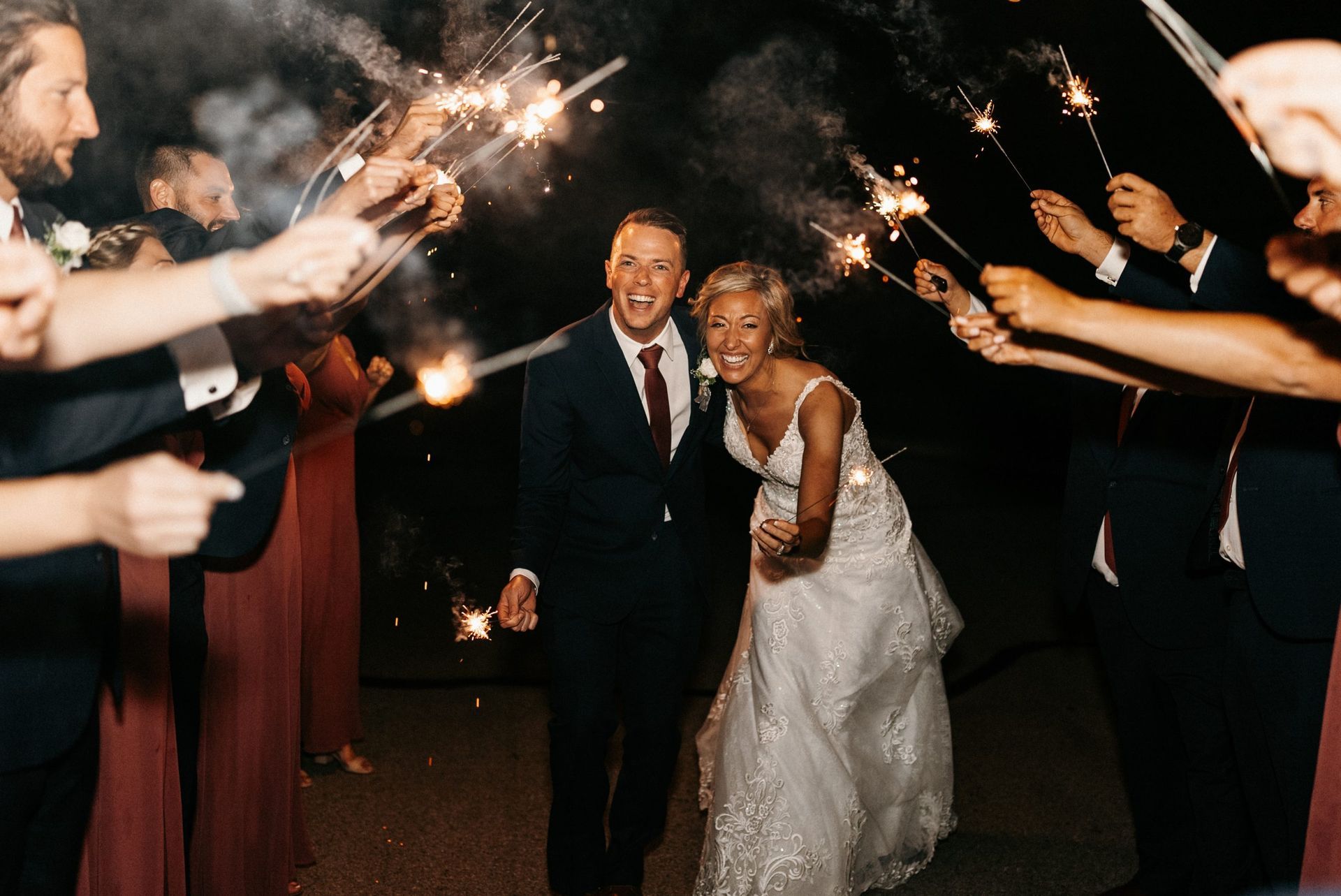 Couple laughs as they walk through sparklers held by wedding guests at night.