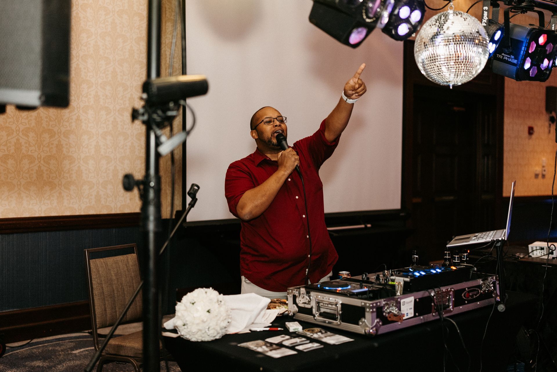 DJ at a party, pointing upward with a microphone, a disco ball and lights above.
