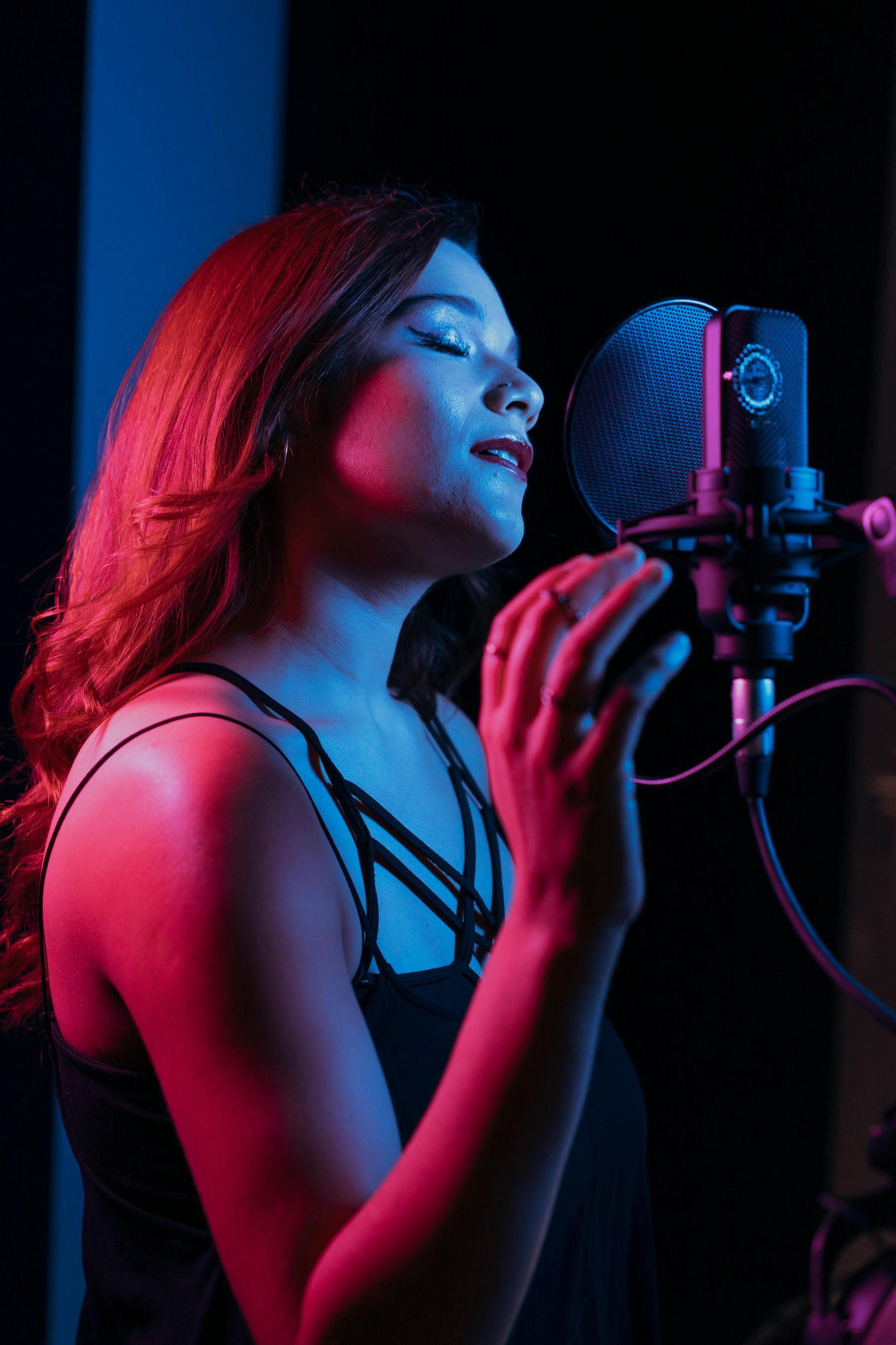 Woman singing into a microphone in a recording studio, lit by red and blue lights.