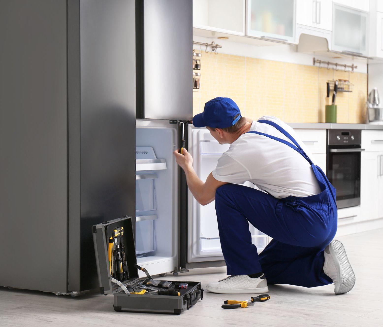 A male technician with a screwdriver is repairing a refrigerator in the kitchen.