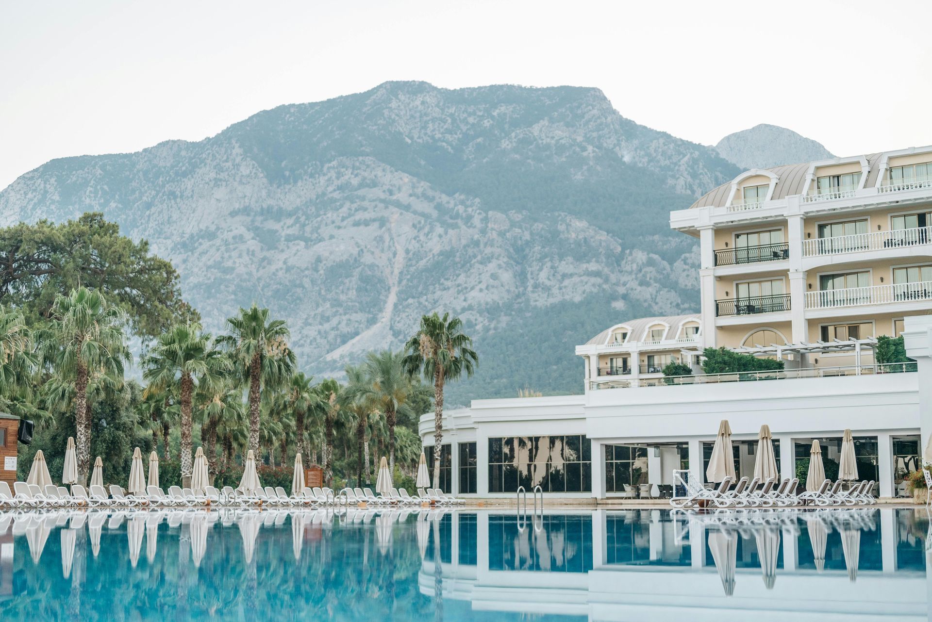 Poolside resort with mountain backdrop. Palm trees, blue water, white building.
