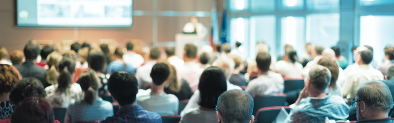 Audience seated in a bright conference hall, facing a presentation screen at the front.