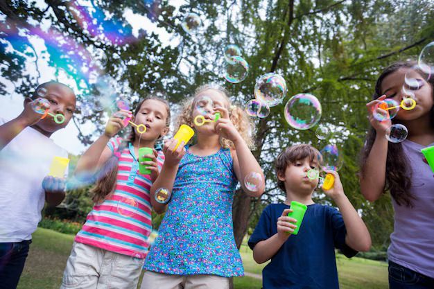 Children blowing bubbles in a park, smiling, with green grass and trees in the background.