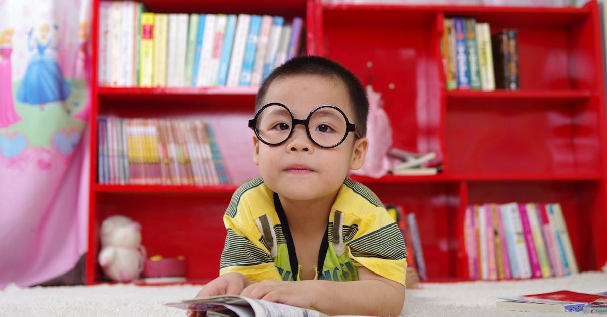 Young child with glasses reading in front of a red bookshelf.