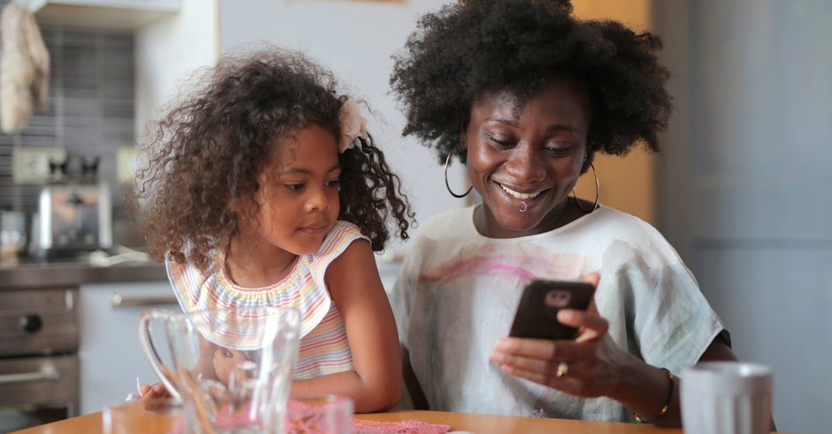 A woman and a little girl are sitting at a table looking at a cell phone.