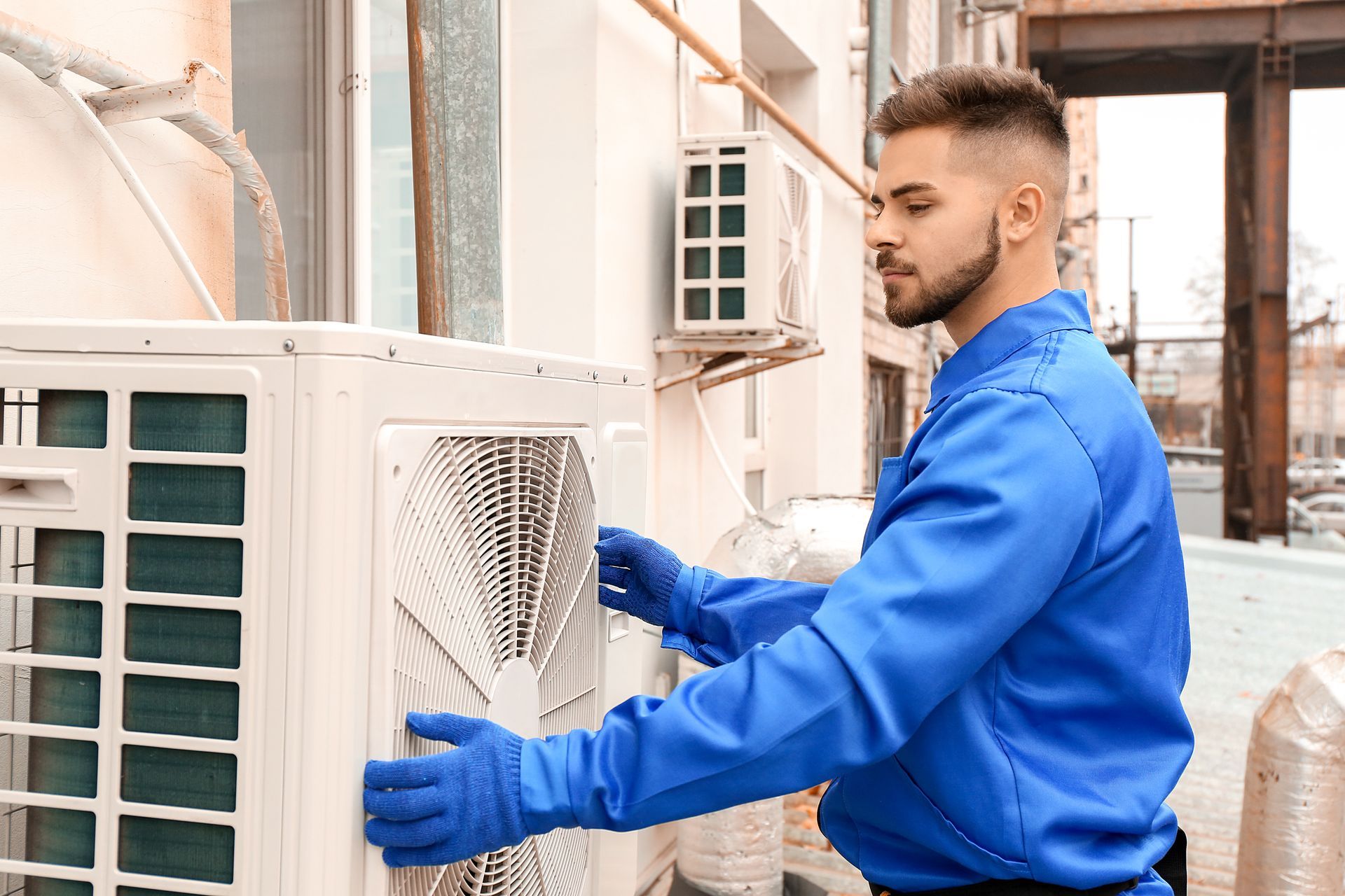 Man in blue uniform installing an AC unit outside a building, wearing gloves.