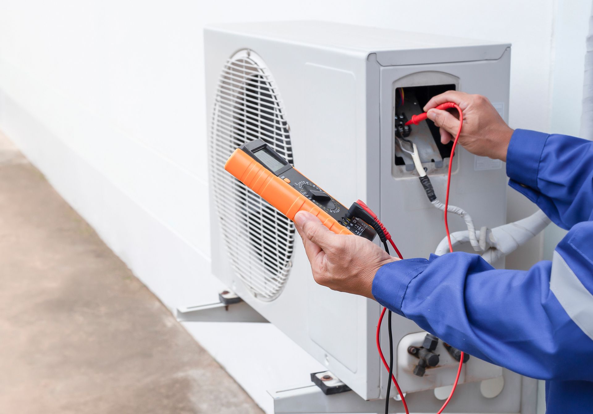 HVAC technician in blue coveralls using a multimeter to test an outdoor air conditioning unit.