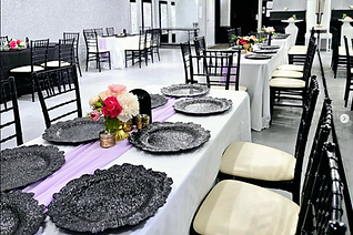 A banquet hall set for an event. Tables are set with floral arrangements, chargers, and chairs. The room is brightly lit.