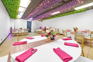 A banquet hall with round tables set with pink linens, floral centerpieces, and gold chairs, decorated with hanging purple and green floral ceiling décor.