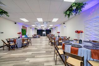 A brightly lit event space with round tables set for a celebration. The tables have blue and orange linens, and wooden chairs.