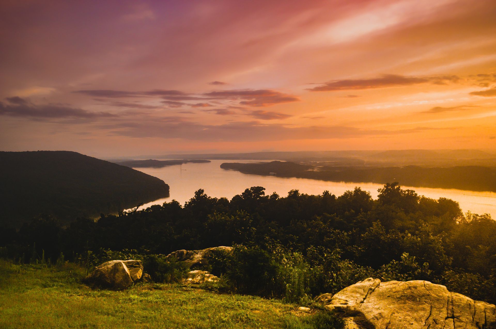 Sunset over a river and hills, with golden sky and grassy foreground rocks