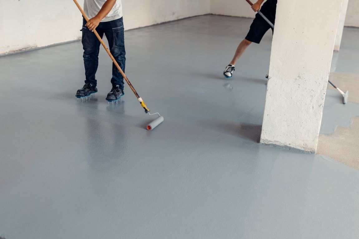 Two people applying gray epoxy flooring with rollers in a room with white walls and a support beam.
