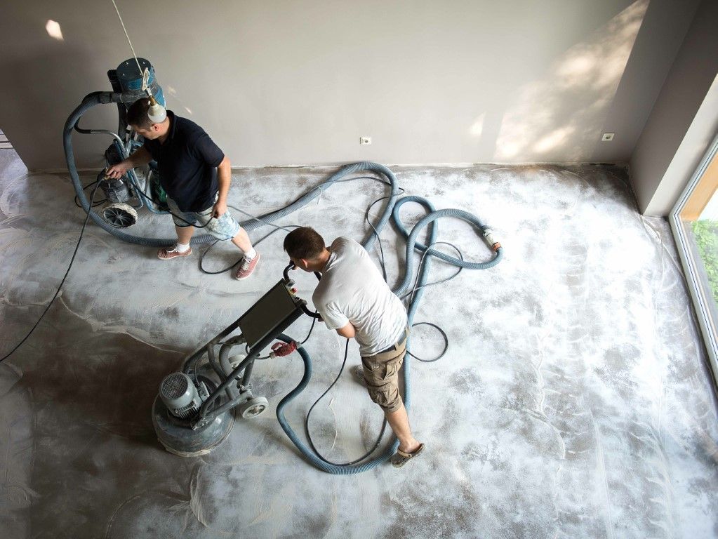 Two men sanding a concrete floor with industrial equipment in a room with light-colored walls.