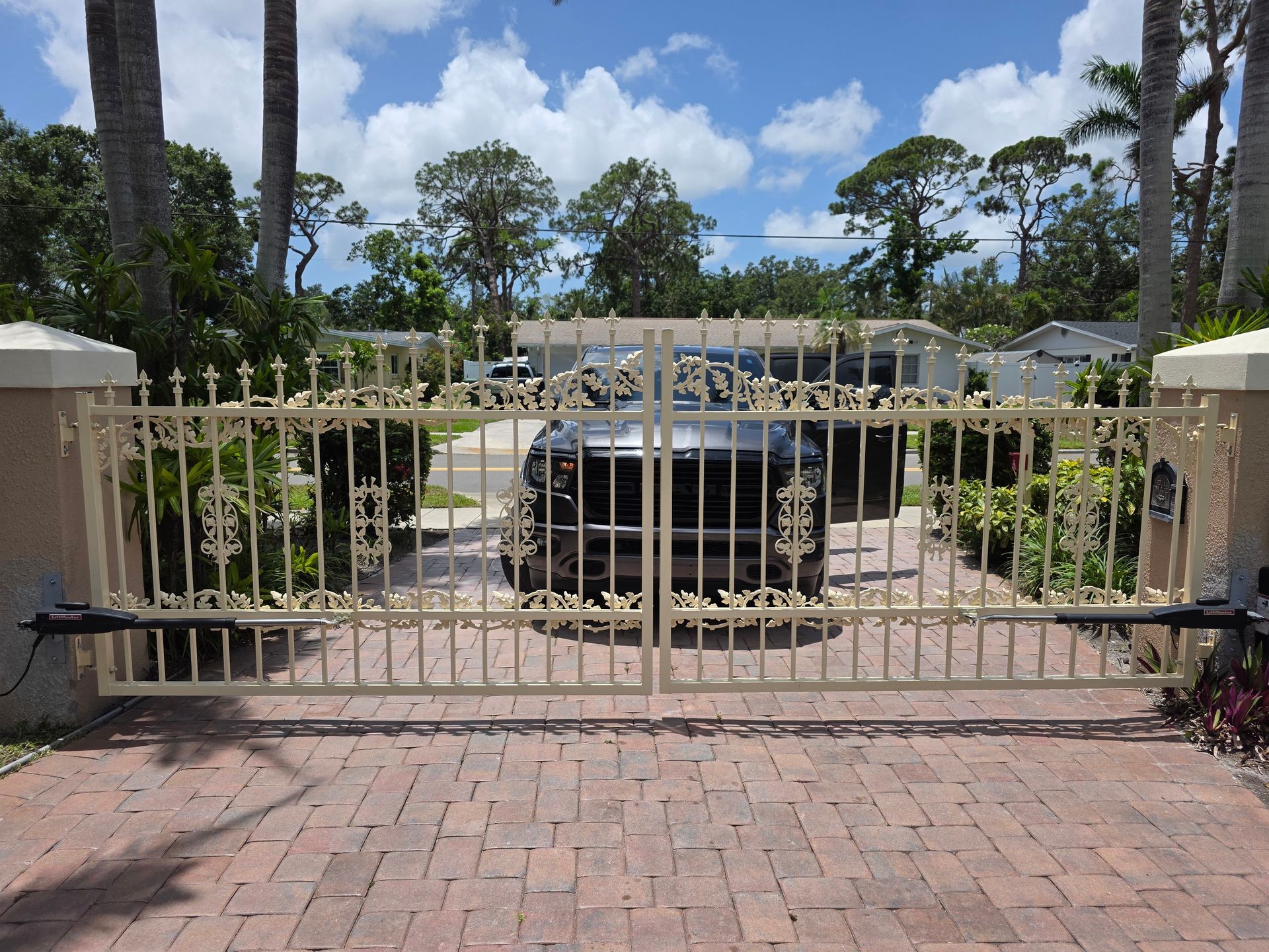 Cream-colored wrought iron driveway gates with a black car parked behind them, set on a brick driveway, on a sunny day.