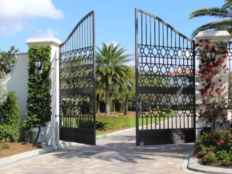 A black gate is open to a driveway with palm trees in the background