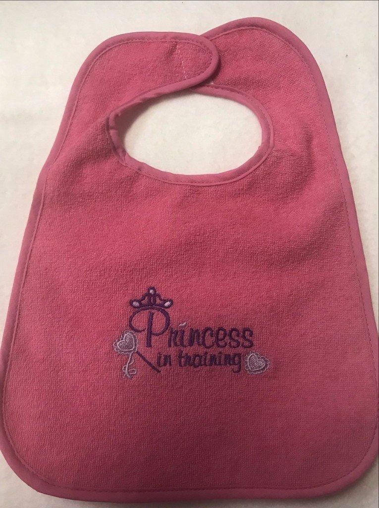 A pink, terrycloth baby bib with an embroidered design featuring a crown and the words 