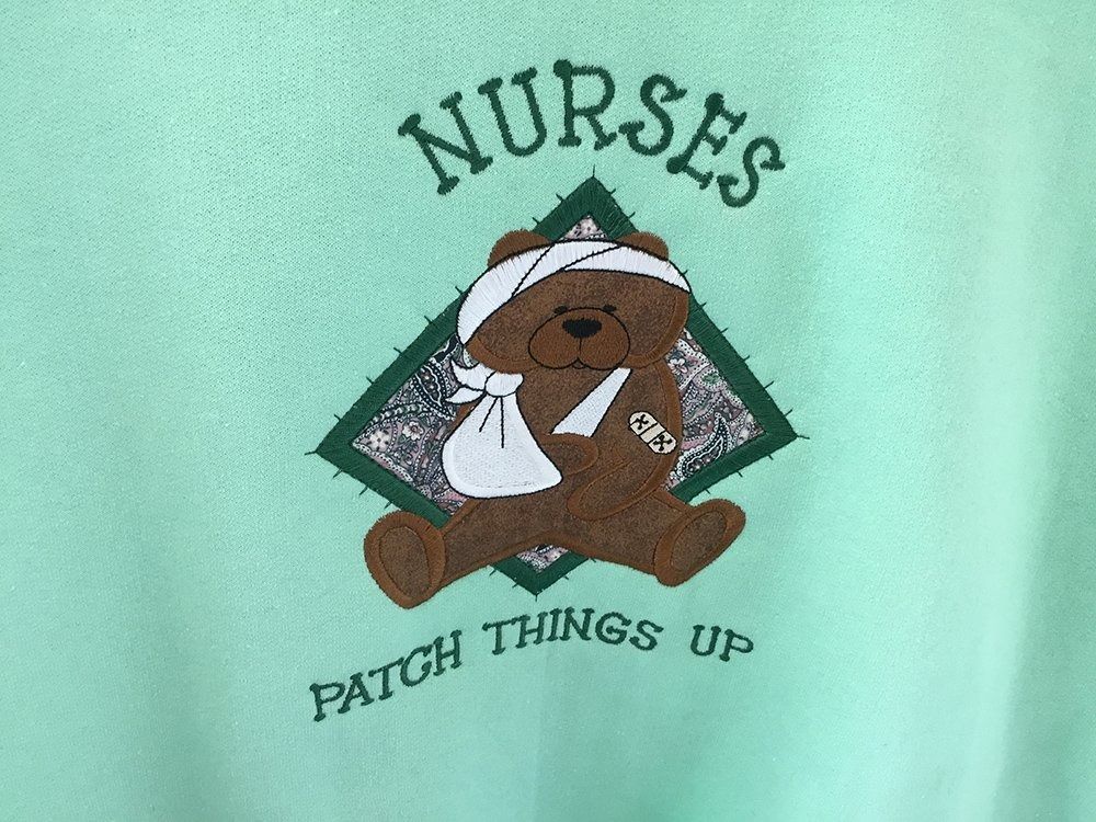 A mint green sweatshirt featuring a brown teddy bear with a bandage on its head, an arm in a sling, and a bandage on its arm.
