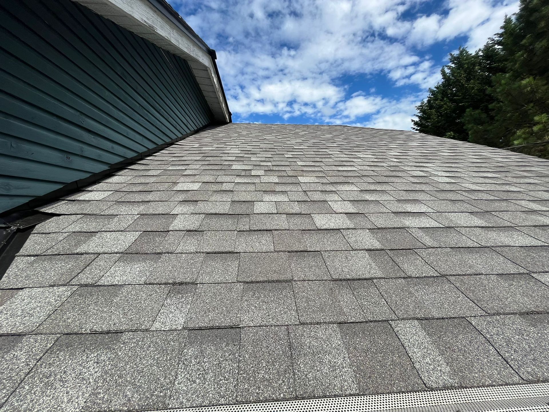 A close up of a roof with shingles and a blue house in the background.