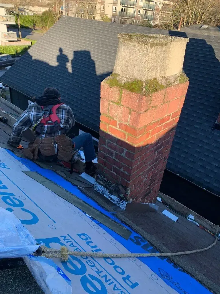 Roofer working on a dark shingle roof near a brick chimney. Sunlight casts a shadow on the chimney and the roof.