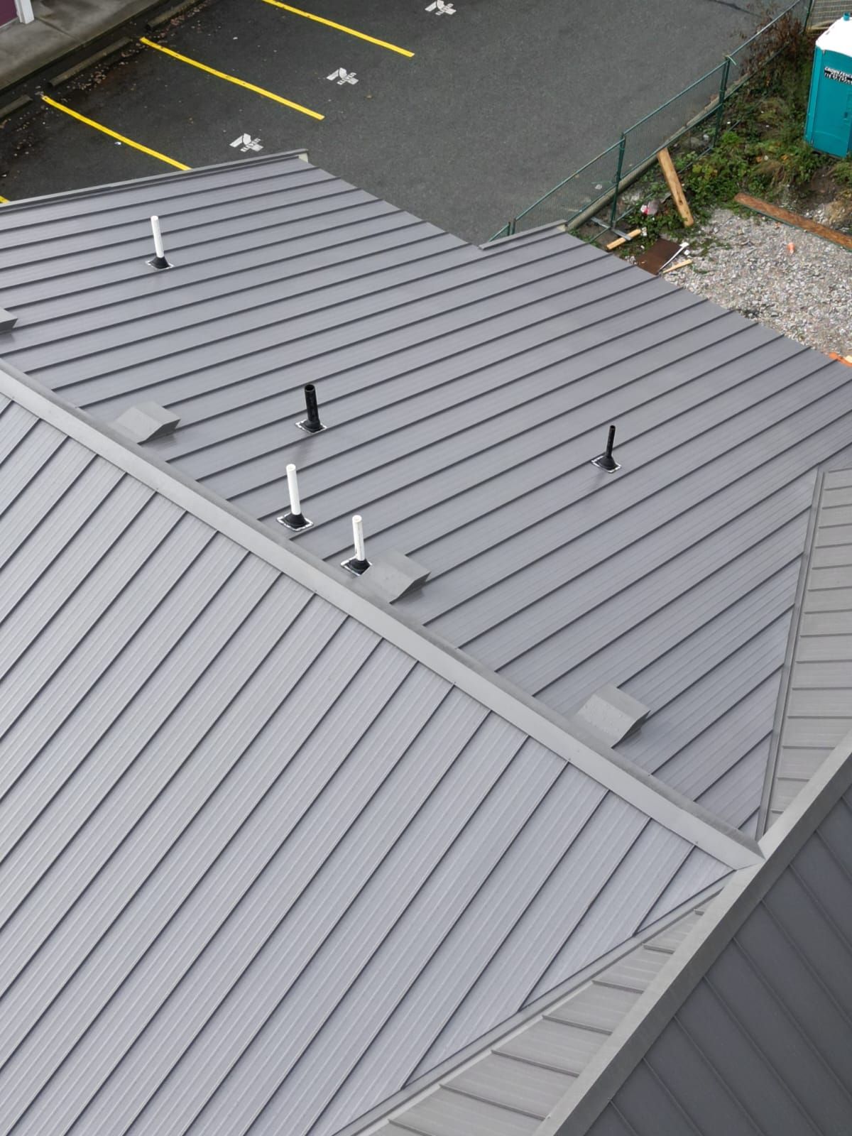 Overhead view of a gray metal roof with vertical lines, vents, and a parking lot in the background.