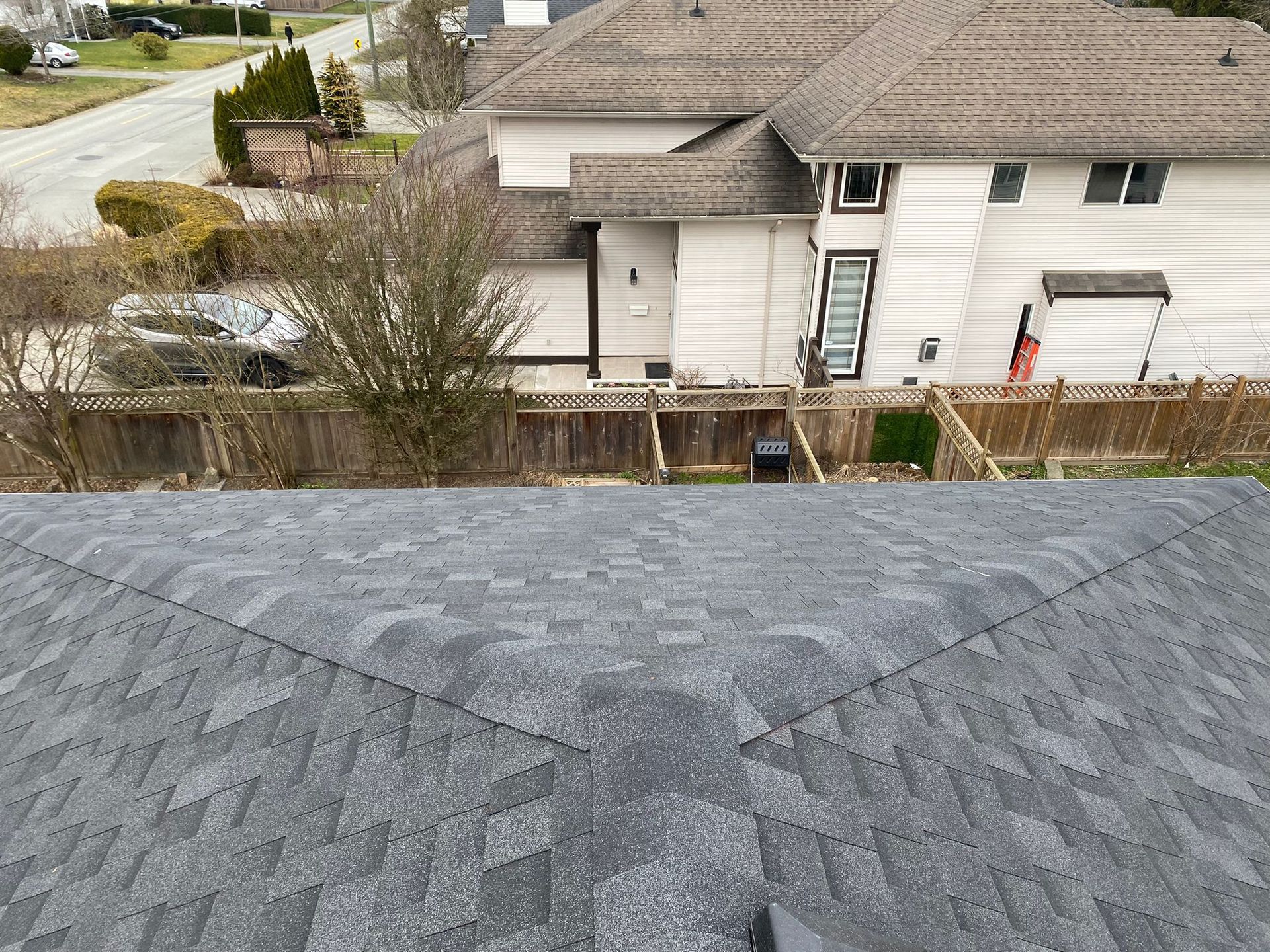 Gray asphalt shingle roof, with a view of a light-colored two-story house with a wooden fence.