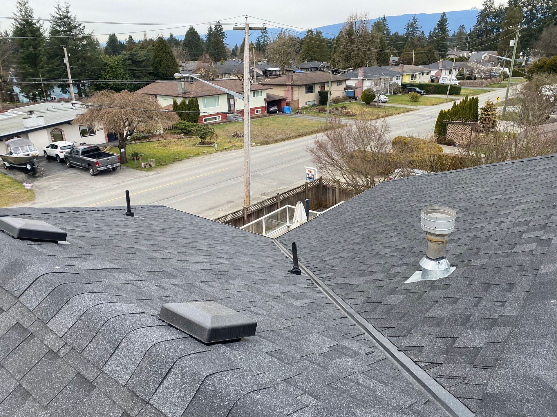 A high-angle view of a gray asphalt shingle roof featuring several vents and a chimney against a suburban street backdrop.
