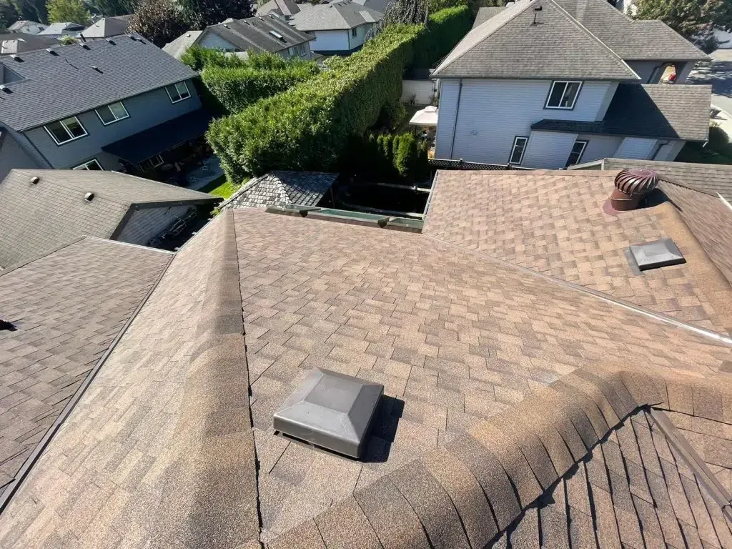 Overhead view of a brown shingled roof with a square vent cap, other homes in the background.