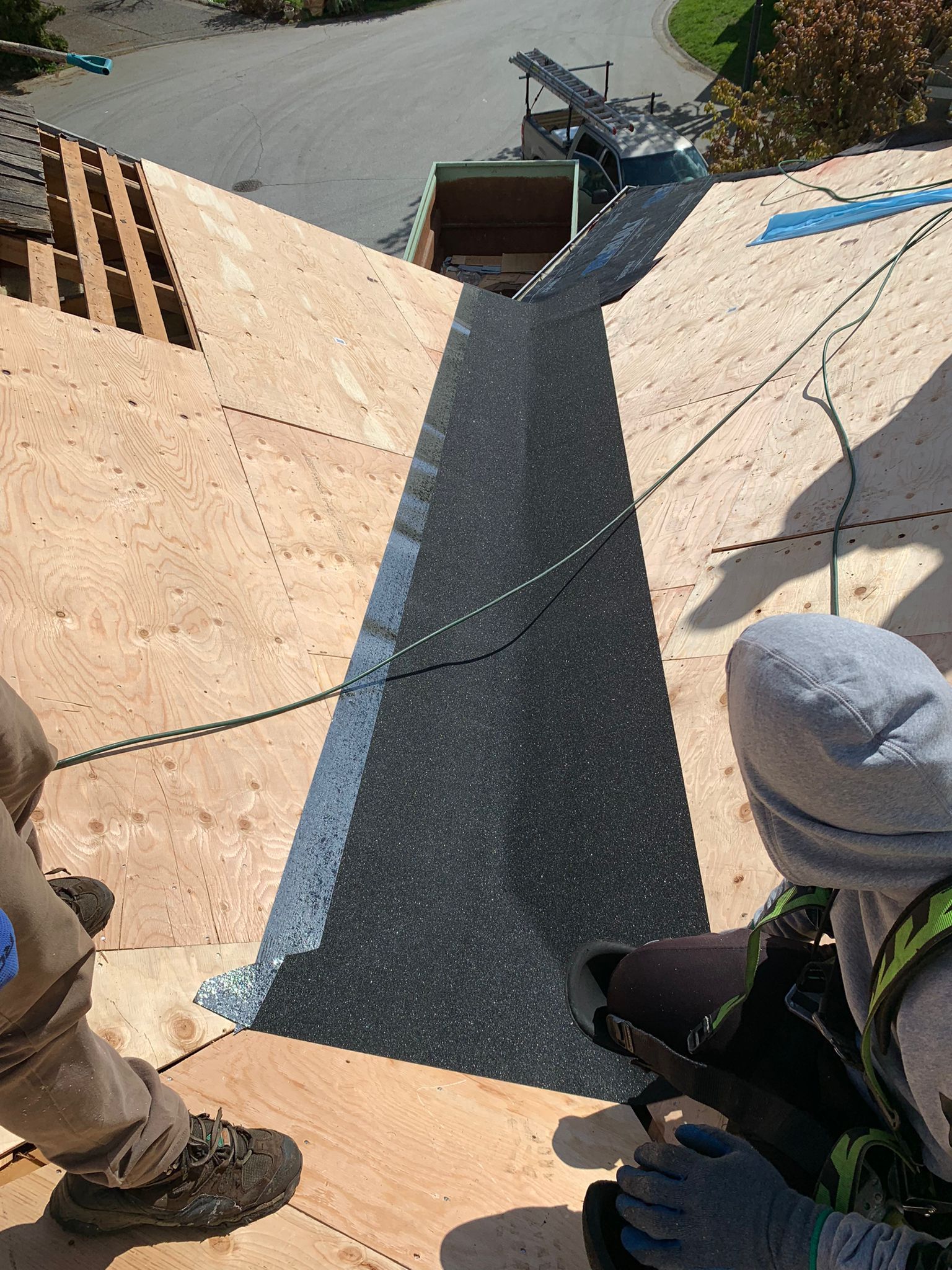 Roofers installing shingles in a roof valley. Black shingle strip installed, wood sheeting visible, daylight.