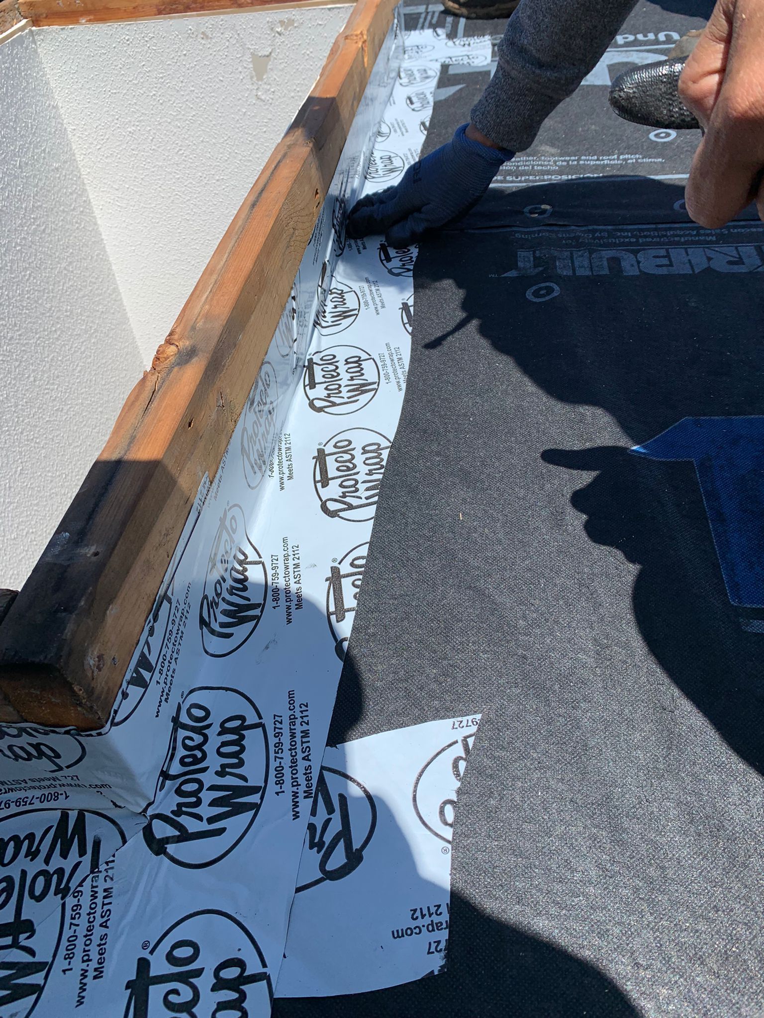 Person applies flashing tape around a wooden frame on a roof, near a black roofing material.