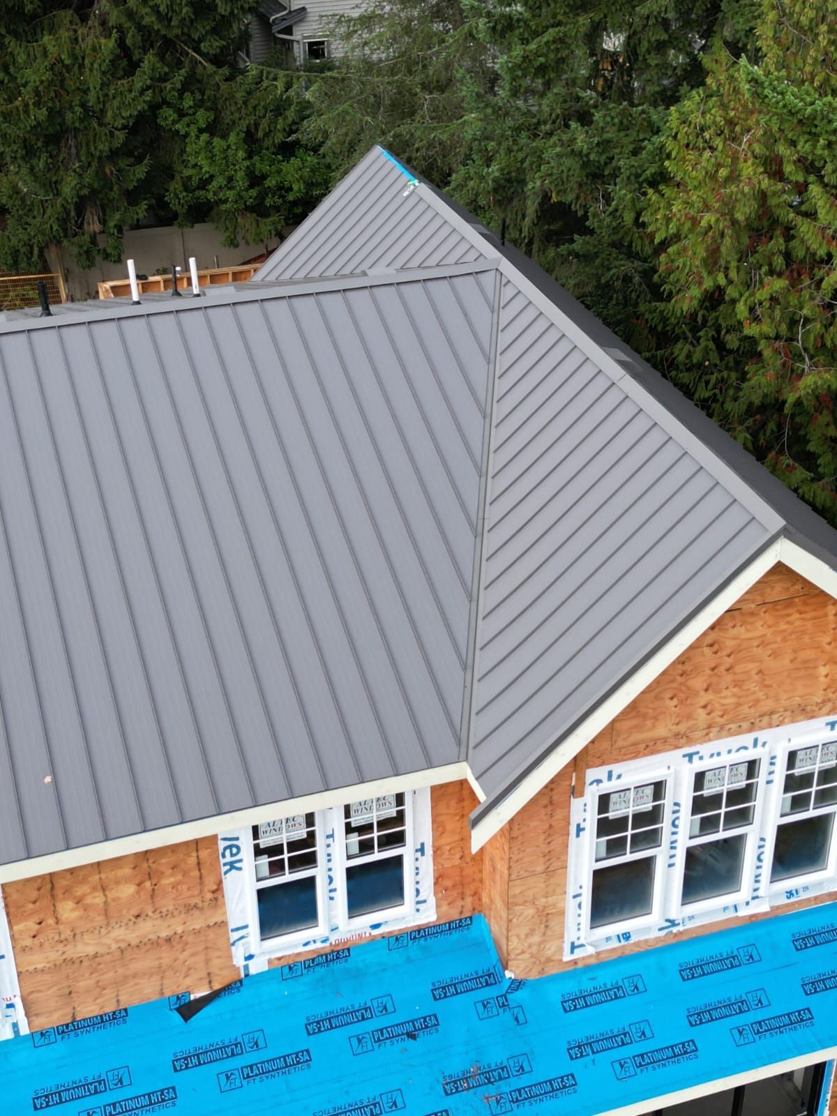 Gray metal roof on a house under construction; windows, wood framing, and trees visible.
