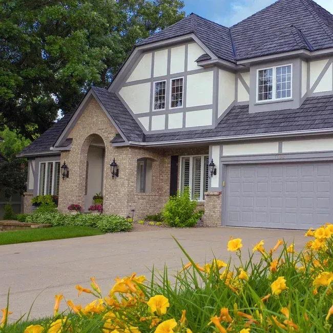 Tudor-style house with gray roof and trim, beige brick, arched entryway, and a driveway with yellow flowers.