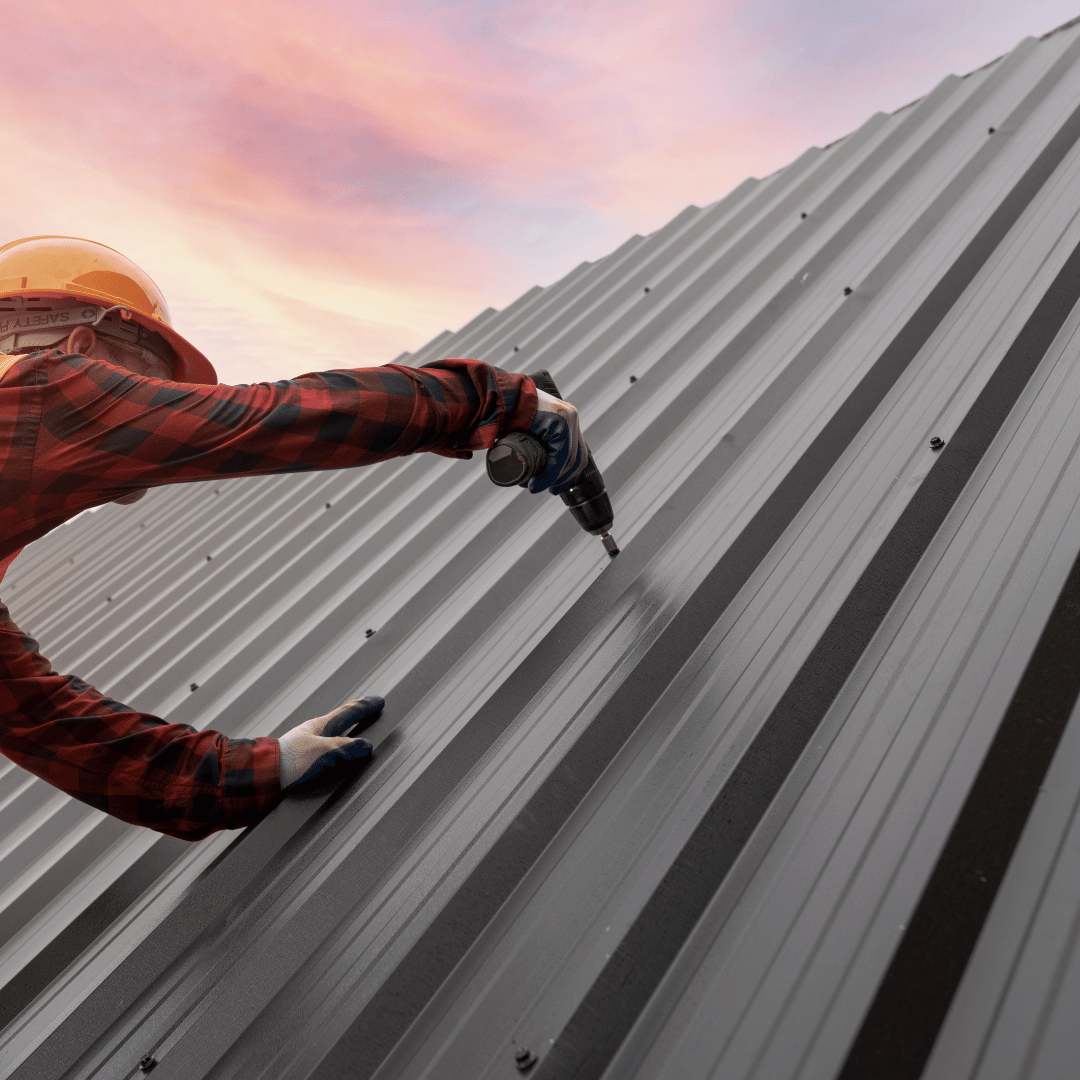 Roofer in hard hat, shirt, and gloves screws metal panels onto a roof at sunset.