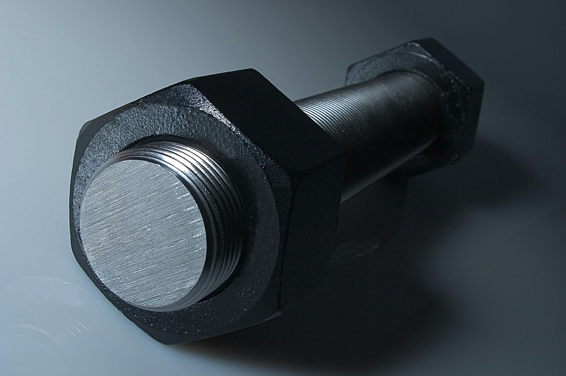 Large, black bolt and nut, close-up against a gray background. The bolt's threads are visible.