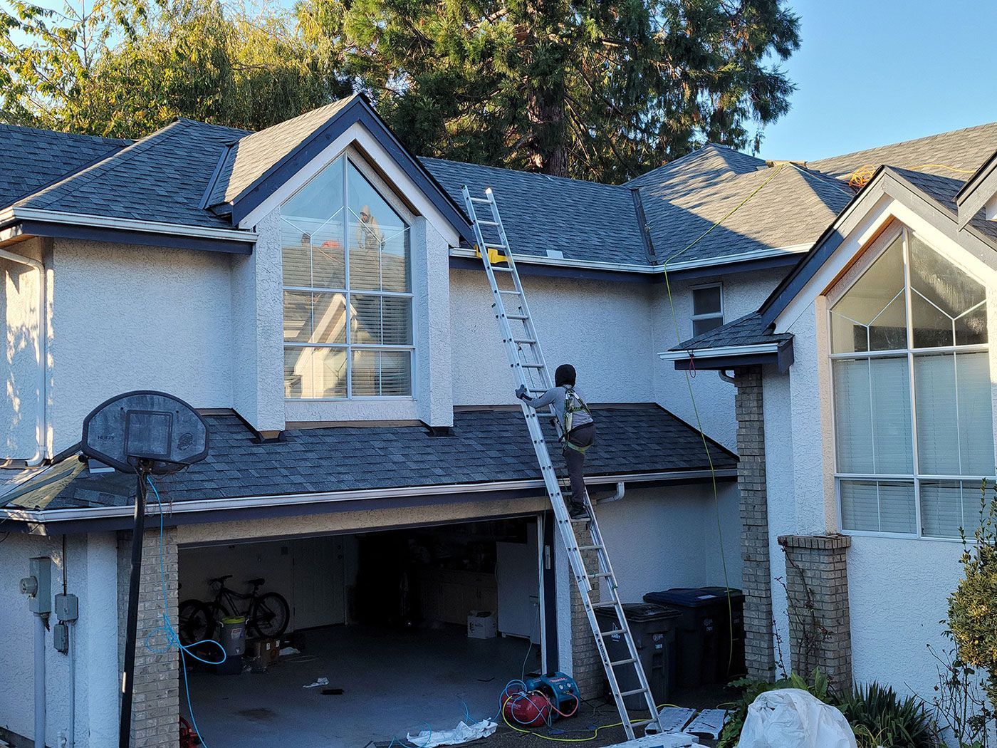 A person on a tall metal ladder works on the roof of a two-story house with a garage and textured light-colored walls.