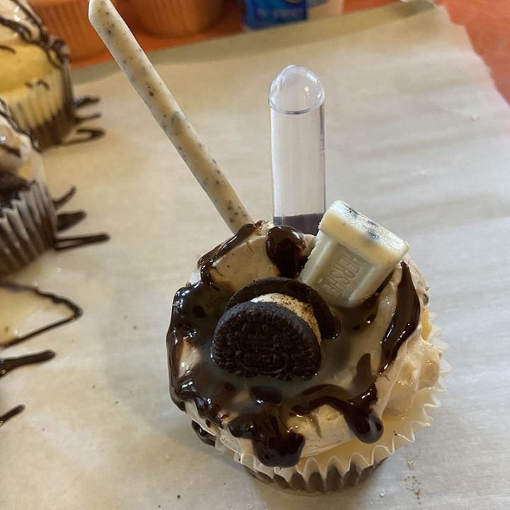 Cupcake topped with chocolate frosting, Oreo, chocolate drizzle, candy stick, and syringe filled with white cream.