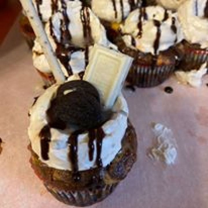 Cupcakes with white frosting, chocolate drizzle, Oreo, and white chocolate in a tray.