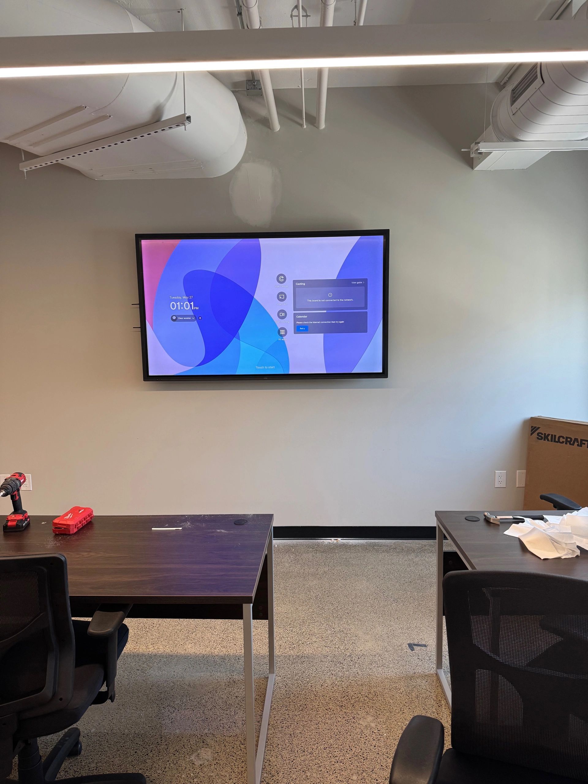 Conference room with wall-mounted TV showing a presentation slide, tables, and chairs in a modern office space.