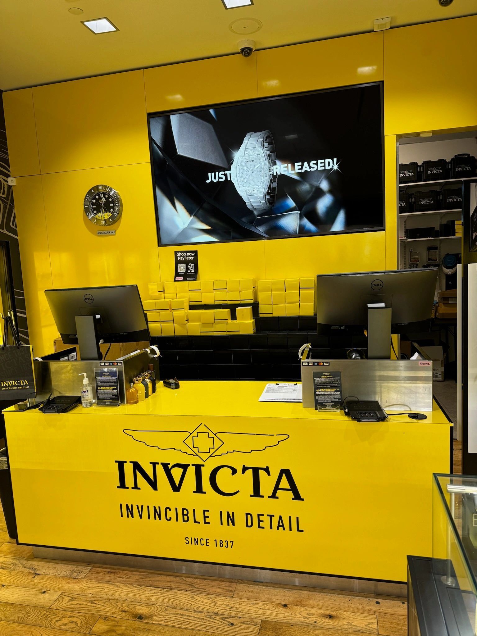 Invicta watch store counter with bright yellow walls and a large display screen overhead