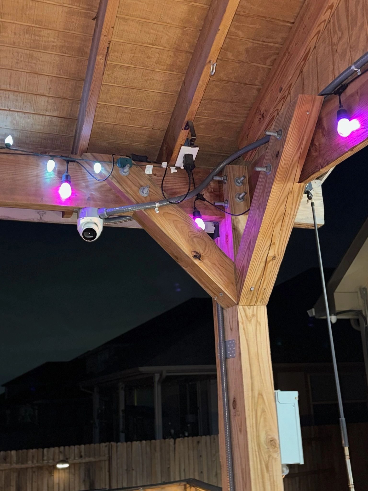 Wooden patio ceiling at night with purple string lights and a mounted security camera.