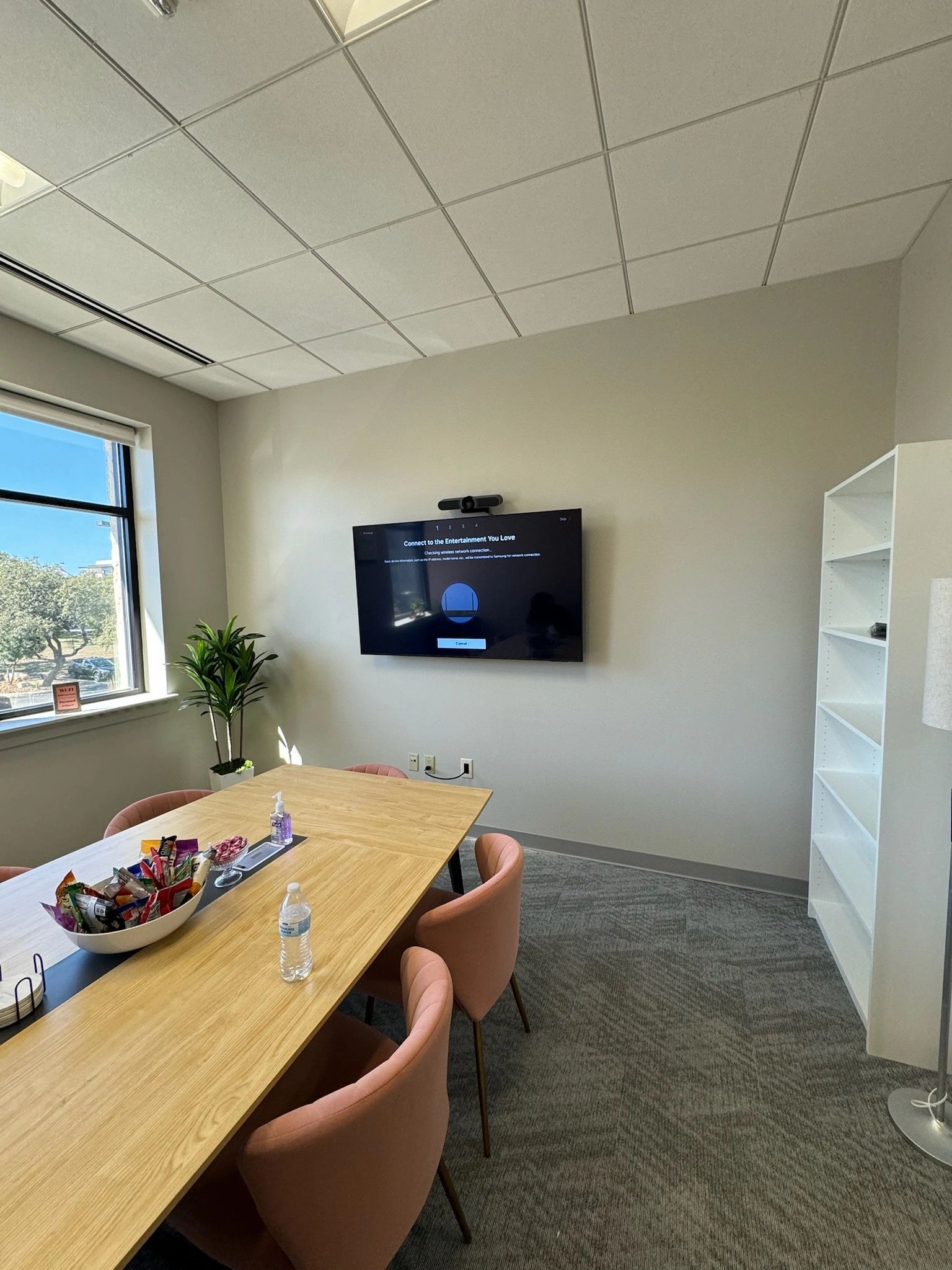 Modern office meeting room with wall-mounted TV, wooden table, pink chairs, and large window.