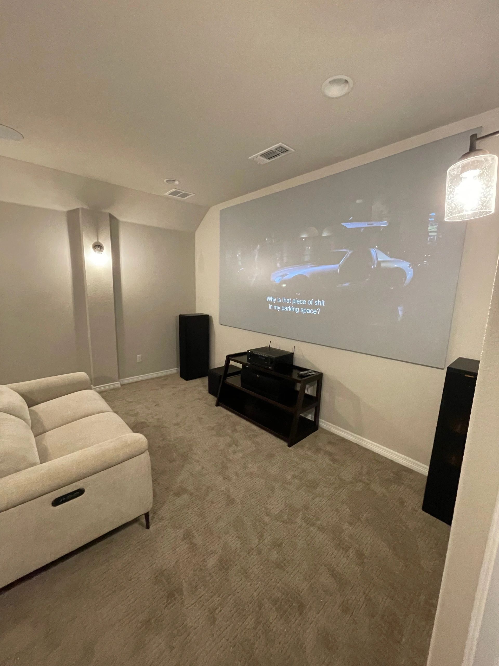 Carpeted home theater room with a projector screen, beige sectional sofa, and black speakers.