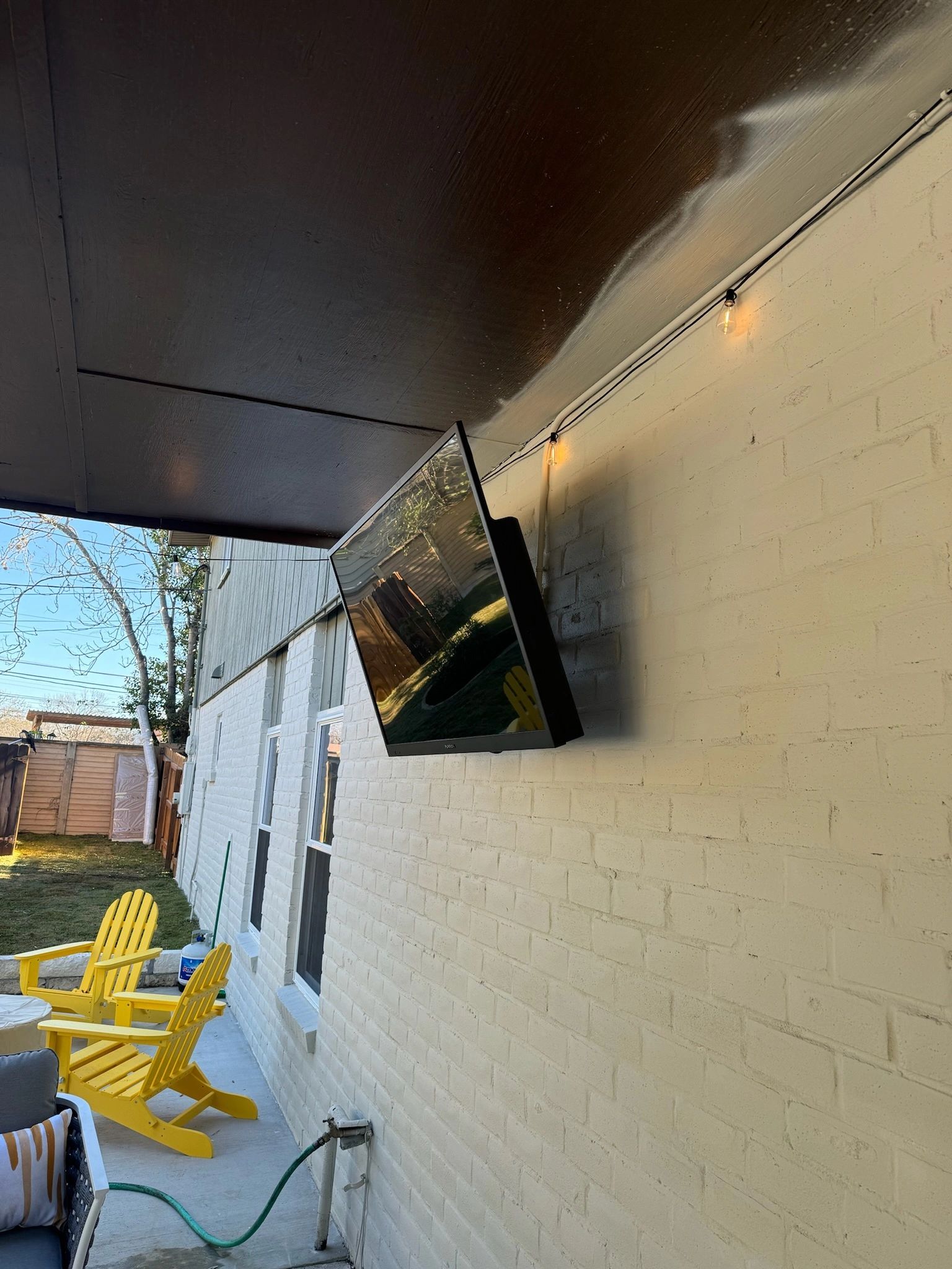 Outdoor patio with yellow chairs, white brick wall, and a mounted TV under a dark awning
