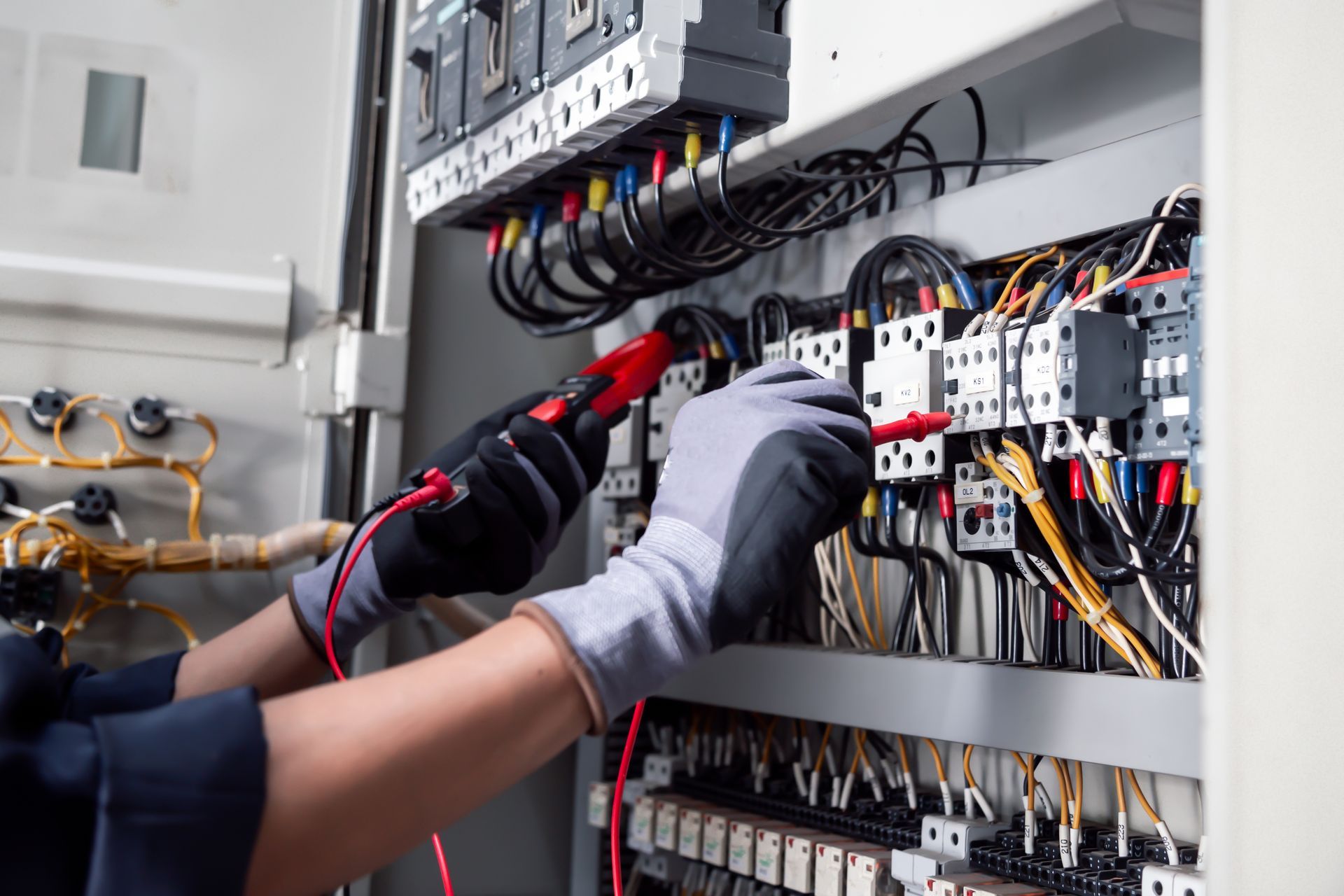 Technician testing electrical wiring inside an open control panel with gloves and a multimeter